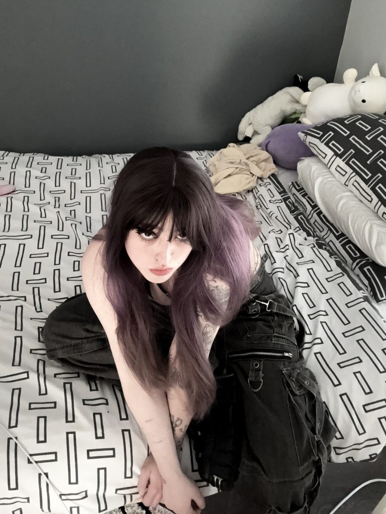 High-angle selfie of Miya on her bed, looking up, showcasing her purple hair, arm tattoos, and black cargo pants.