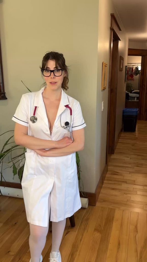 Full-length portrait of Nova Nickels standing in a house hallway, wearing glasses and a short-sleeved white medical lab coat or nurse uniform with a stethoscope around her neck.