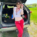 Full-length shot of Nova Nickels leaning against the open trunk of a gray SUV parked outside on grass, wearing glasses, a white crop top, red medical scrub pants, and a stethoscope.