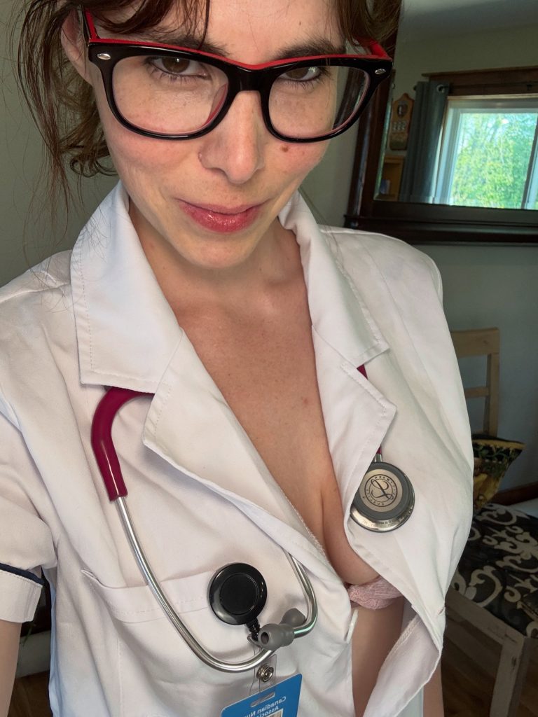Close-up selfie of Nova Nickels wearing black-rimmed glasses, a stethoscope, and an open white medical coat over a pink lace bra.