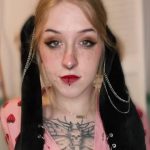 Close-up portrait of Paleseafoam, a young woman with blonde hair, green eyes, and distinct facial piercings, wearing a pink strawberry-patterned shirt and black plush rabbit ears adorned with silver chains.