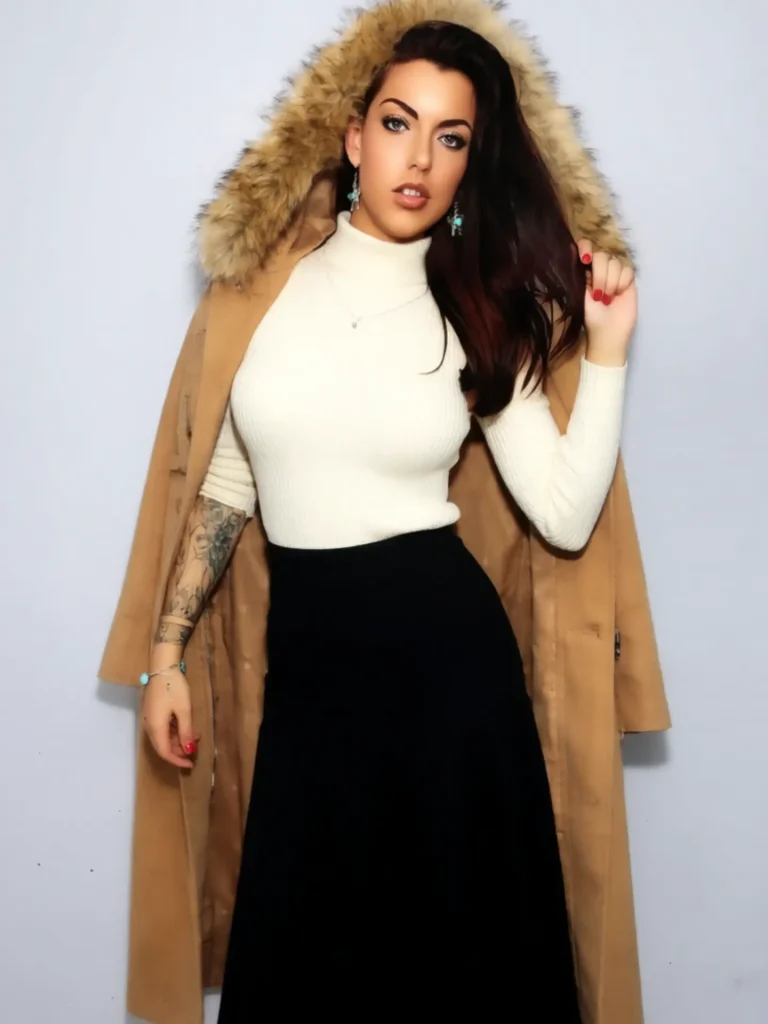 South African OnlyFans personality Skye Blye posing against a white wall, wearing a white turtleneck and a tan coat draped over her shoulders.