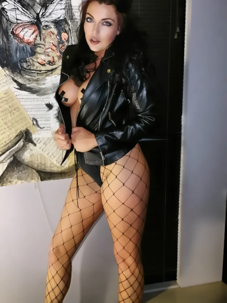 Skye Blye in a leather jacket and fishnet stockings, standing next to a butterfly art piece.