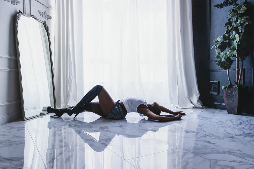 Nigerian OnlyFans model Tayy Knoxx posing on a polished marble floor in denim shorts and thigh-high boots in front of bright sheer curtains.