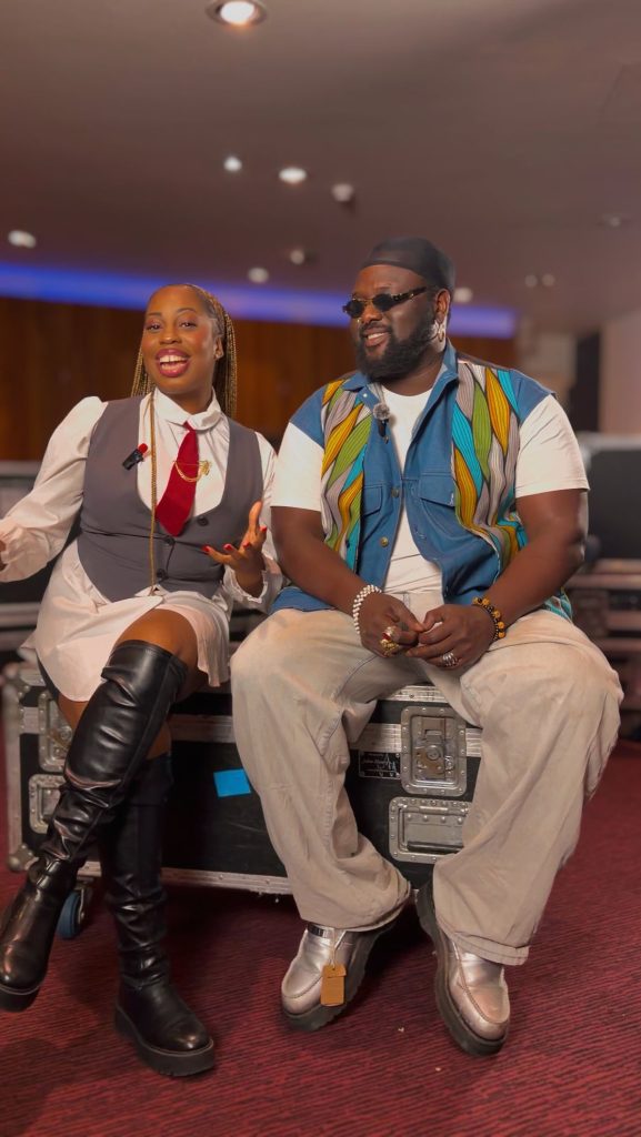 Nigerian OnlyFans creator Tommy Kuti (right), wearing a patterned denim vest over a white t-shirt and loose light-colored jeans, sits on a black equipment road case next to a woman in a grey waistcoat and red tie, both smiling.