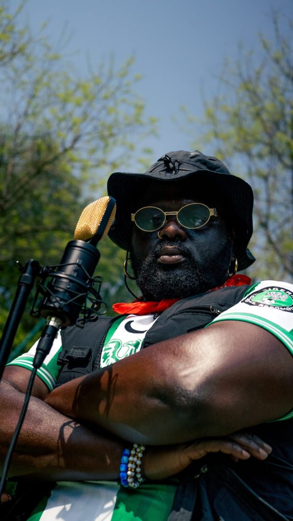 A close-up, low-angle shot of Nigerian OnlyFans creator Tommy Kuti, wearing a black bucket hat, round sunglasses, and a black vest over a green patterned shirt, posing with arms crossed outdoors in front of a golden microphone against a background of green trees.