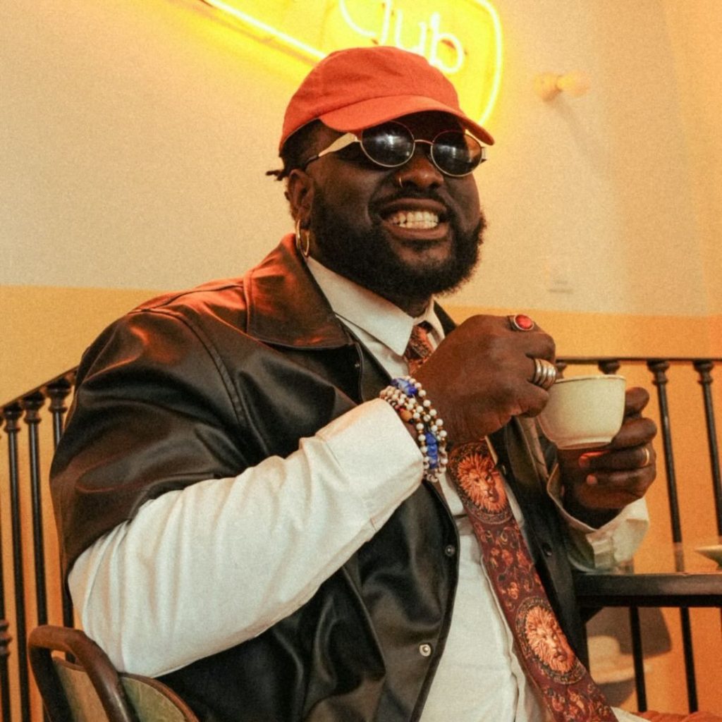 A close-up portrait of Nigerian OnlyFans creator Tommy Kuti, smiling broadly and holding a small white coffee cup, wearing an orange baseball cap, round sunglasses, and a black leather shirt over a white collared shirt with a patterned tie.