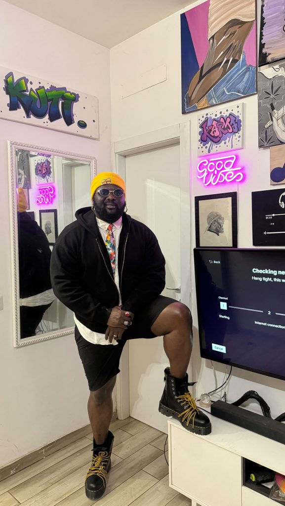 A full-length indoor portrait of Nigerian OnlyFans creator Tommy Kuti, wearing a yellow beanie, a black open hoodie over a white shirt and patterned tie, black shorts, and black combat boots, posing with one leg raised on a small white cabinet in a room decorated with graffiti art and a pink neon sign.