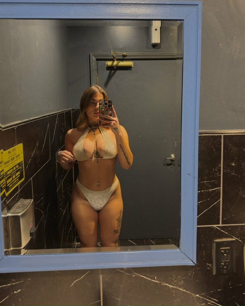A bathroom mirror selfie of Lilbadblue posing in a white, embellished halter-neck bikini set.