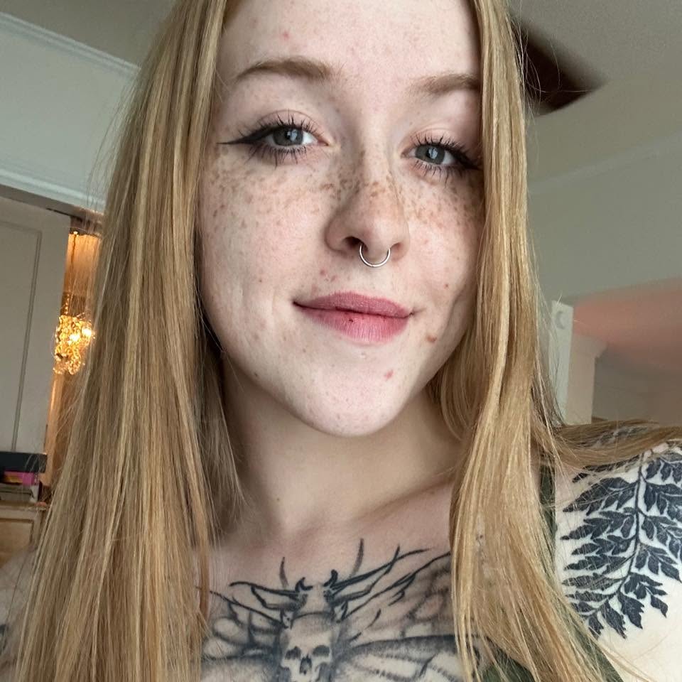 Close-up selfie of Paleseafoam, a smiling young woman with long blonde hair, prominent freckles across her nose and cheeks, and a silver septum piercing. She has a large skull and moth tattoo visible on her chest, as well as a leafy fern tattoo on her shoulder.