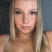 Close-up portrait of a blonde woman named Bellaabuns looking directly at the camera, wearing a thin gold necklace with small pendants.