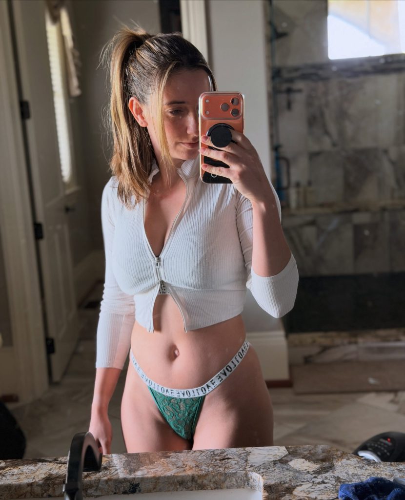 Woman taking a bathroom mirror selfie in a white zip-up crop top and green lace underwear, featured in a guide to the best OnlyFans to buy subscriptions for exclusive daily sets.