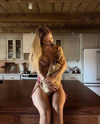 Blonde fitness creator Daisy Keech sitting on a wooden kitchen island holding a small dog, styled for Muscular Women OnlyFans creators.