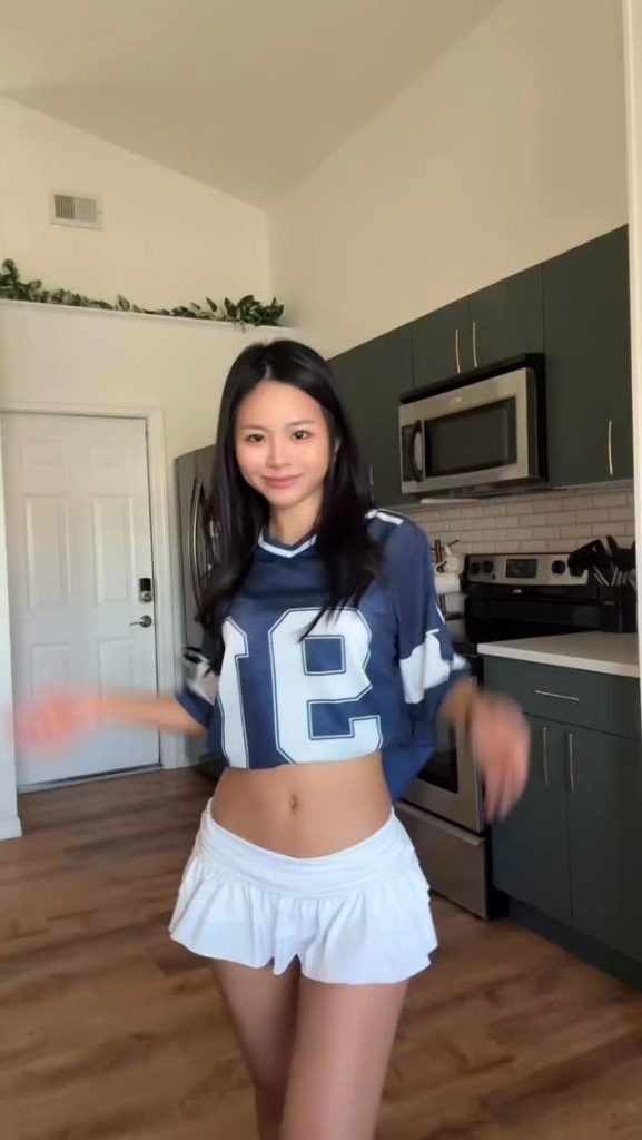 Hanna Zuki posing in a kitchen wearing a cropped navy football jersey and a white pleated mini skirt.