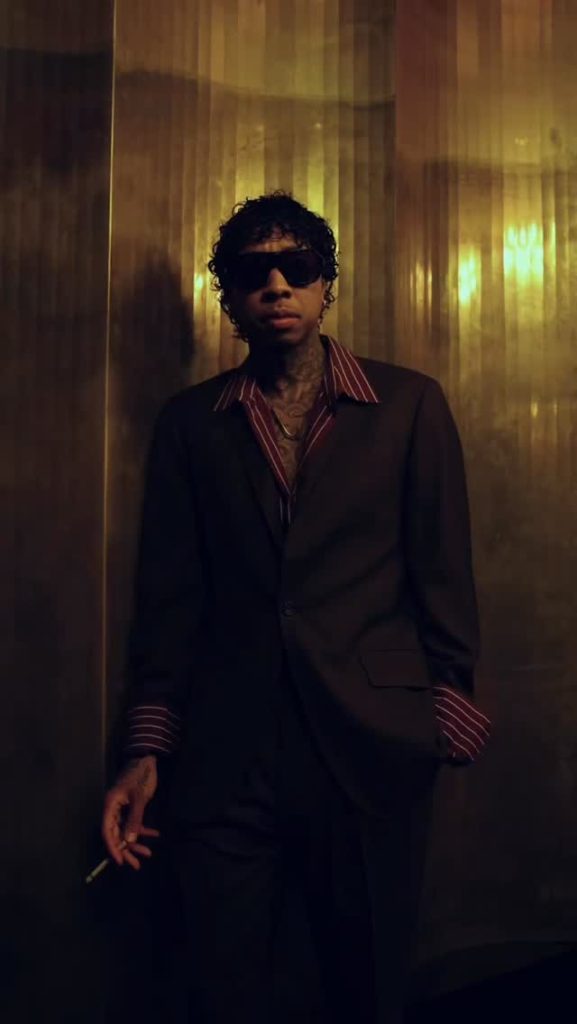 Tyga Rapper standing in moody lighting wearing a dark suit, red striped shirt, and sunglasses while holding a cigarette.