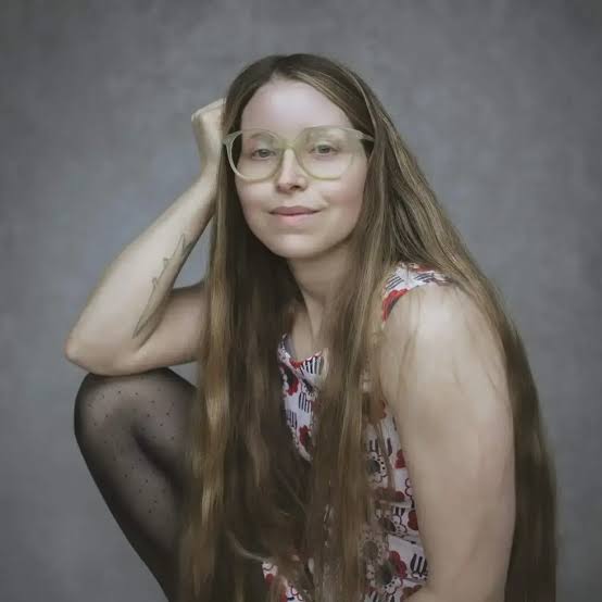 Jessie Cave Harry Potter actress posing with long straight hair and light-framed glasses in a simple portrait shot.