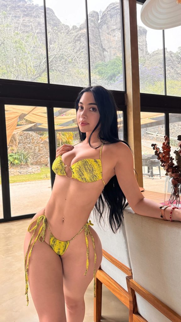Jailyne Ojeda wearing a yellow snakeskin-print string bikini indoors with large windows and scenic mountains in the background