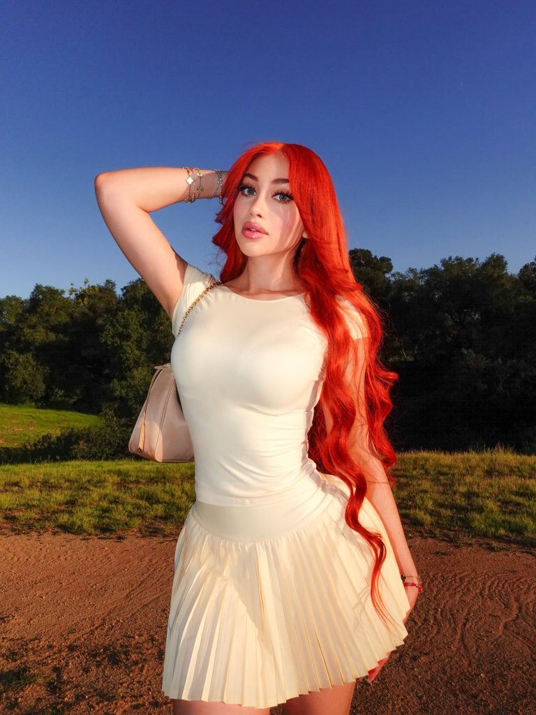 Jailyne Ojeda with long bright red hair posing outdoors in a white top and pleated skirt