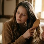 Jessie Cave from the Harry Potter film series wearing a cozy sweater and using a wooden hairbrush at home.