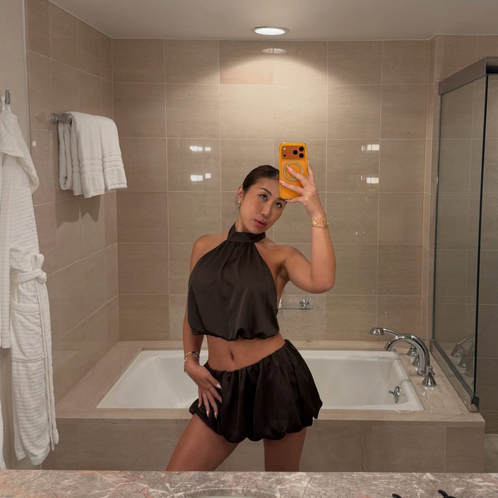 Nicole Doshi, an Asian American OnlyFans creator, taking a mirror selfie in a bathroom with a large bathtub, wearing a brown satin halter top and matching bubble shorts.