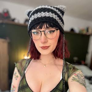 Portrait of a Femboy OnlyFans creator with dark red hair wearing a striped cat-ear beanie and glasses.