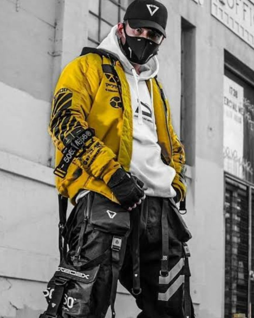 A person posing outdoors in a futuristic yellow and black cyberpunk techwear jacket and face mask, demonstrating top cosplay trends in streetwear fusion