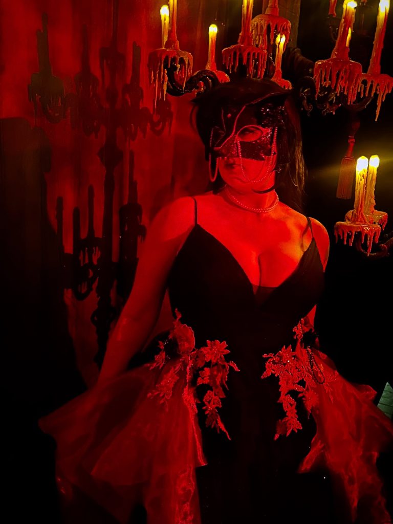 Top Convention Cosplayers featuring a masquerade character wearing a ornate lace mask and black dress under atmospheric red lighting with dripping candles