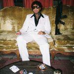 Tyga Rapper sitting on a velvet couch in a white suit and dark sunglasses next to a coffee table with playing cards and casino chips.