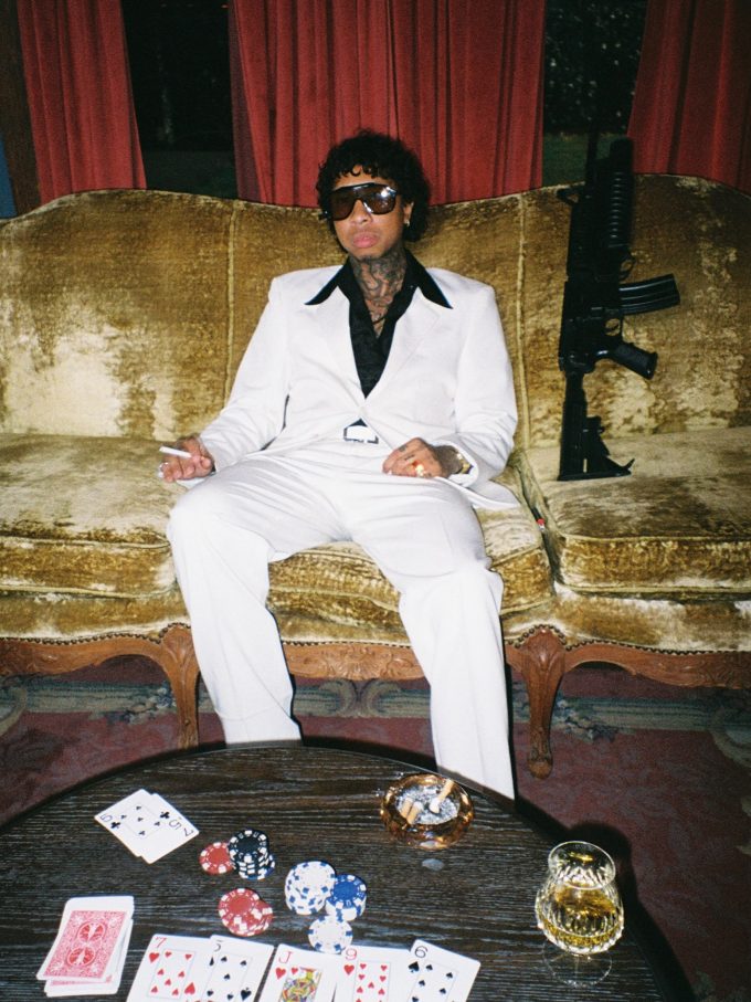 Tyga Rapper sitting on a velvet couch in a white suit and dark sunglasses next to a coffee table with playing cards and casino chips.