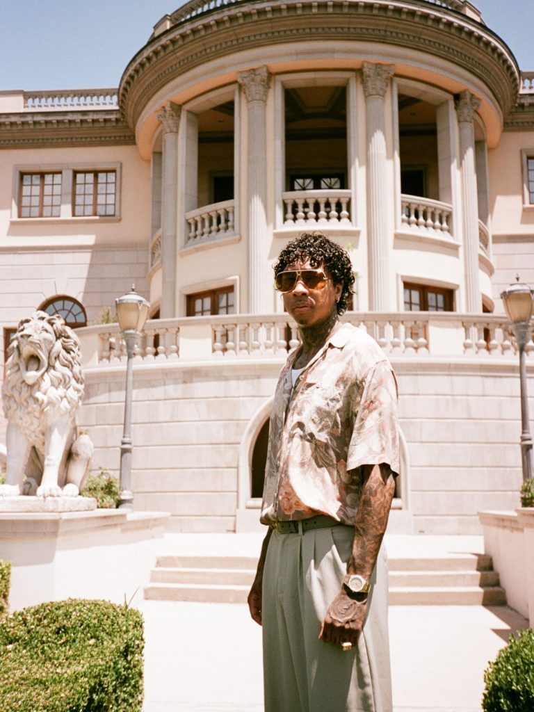 Tyga Rapper posing outdoors in sunglasses and a short-sleeved patterned shirt in front of a luxury mansion.