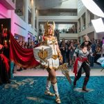Top Convention Cosplayers posing together at a fan convention including She-Ra, an Assassin's Creed assassin, a gothic masquerade character, and a traditional Chinese fantasy character