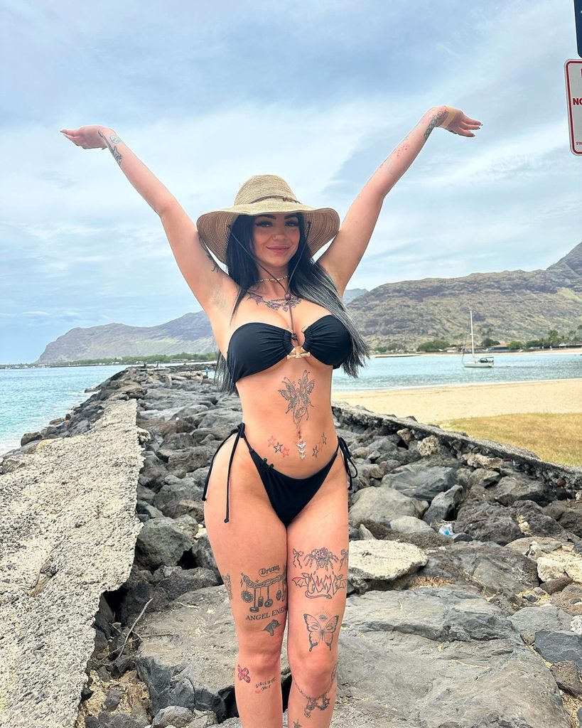Smiling, heavily tattooed OnlyFans influencer in a straw hat and black bikini poses with arms up on a rocky breakwater by the ocean.
