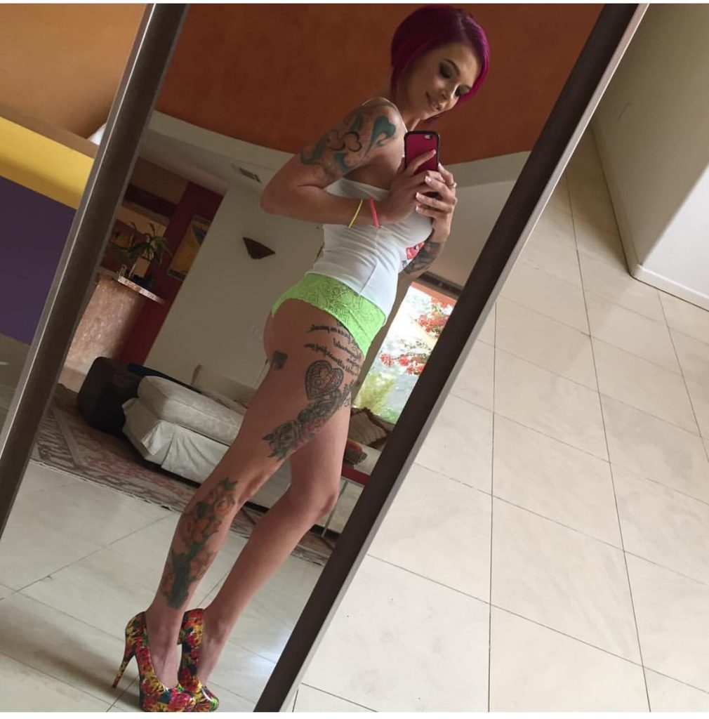 Mirror selfie of tattooed OnlyFans creator Anna Bell Peaks wearing a white top and patterned heels, showing off extensive leg and arm tattoos.