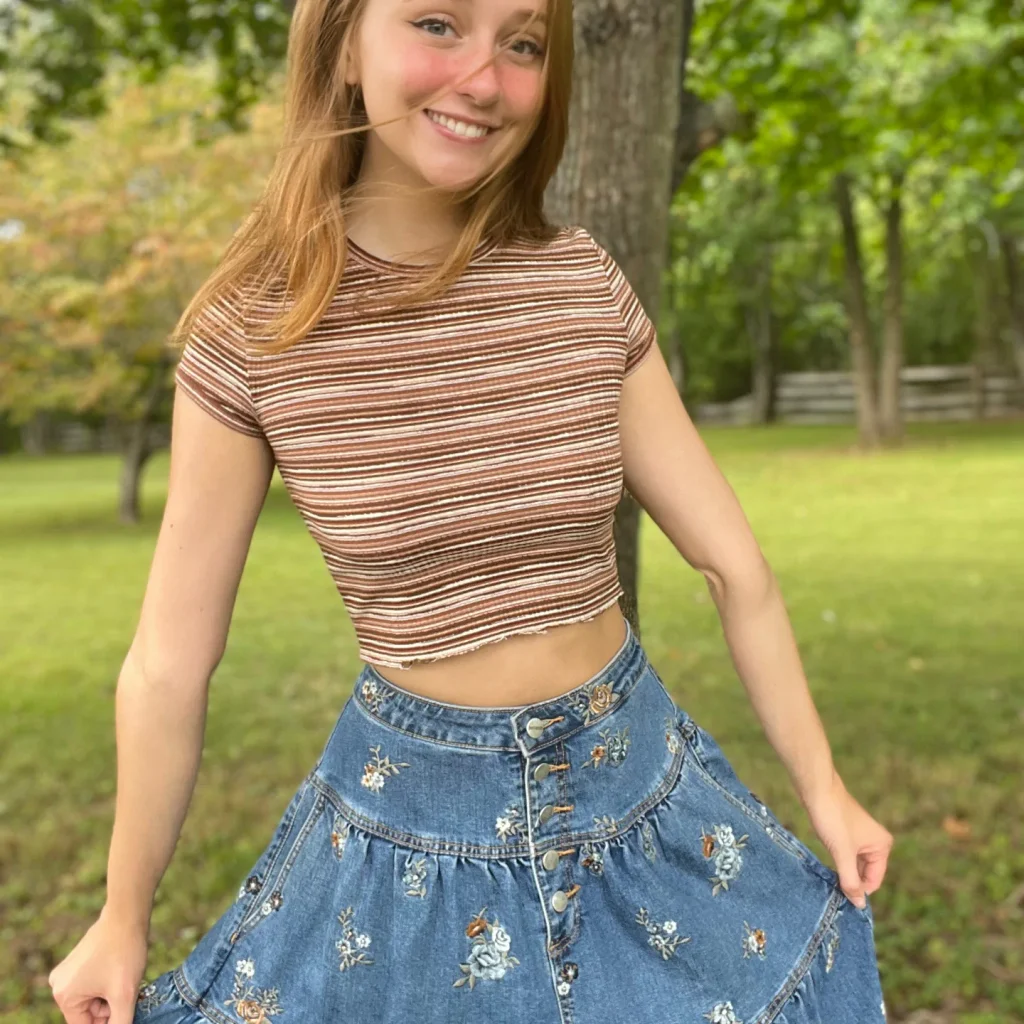 A smiling redhead woman outdoors, wearing a brown striped crop top and a floral denim skirt, styled for a teacher onlyfans profile.