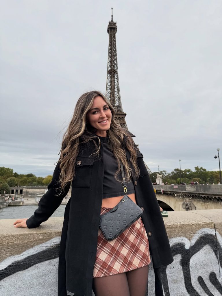 A portrait of a smiling woman posing in Paris near the Eiffel Tower, styled in a plaid skirt and black jacket reminiscent of the Brat OnlyFans fashion trend