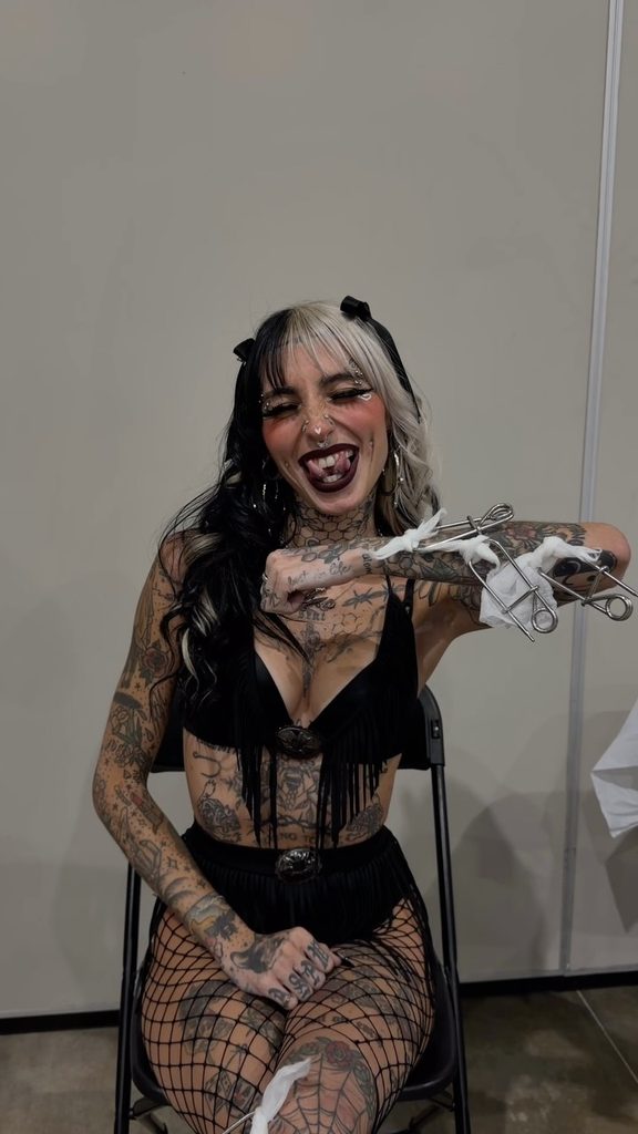 Tattooed OnlyFans model Cruella Morgan with split-dyed black and white hair smiling in a black outfit and fishnets.