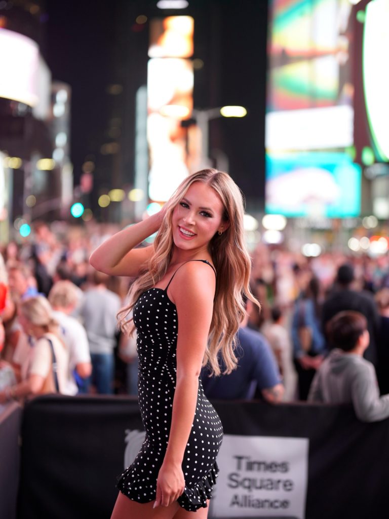 A smiling blonde teacher onlyfans creator wearing a black polka dot dress while posing in Times Square at night with a blurred crowd and city lights in the background