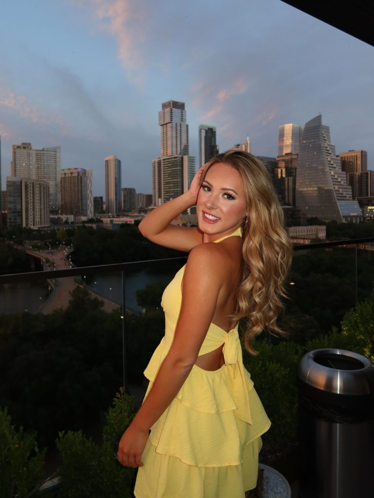 A blonde teacher onlyfans star wearing a yellow dress and posing on a rooftop terrace overlooking a city skyline at dusk