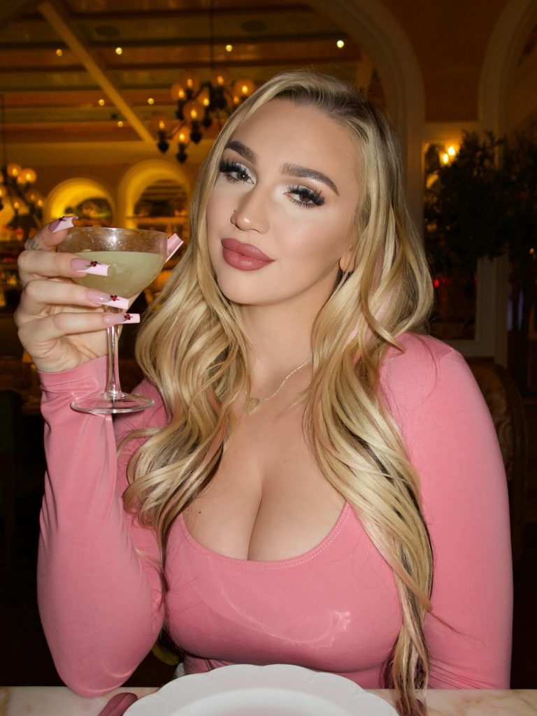 A blonde OnlyFans model in a fitted pink top raising a cocktail glass at an elegant restaurant dining table.