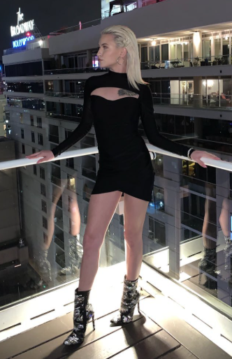 A stylish blonde woman with short hair wears a black long-sleeve mini-dress and sequined ankle boots, posing on a city balcony at night with a "Broadway Hollywood" sign in the background. Her fashion-forward style and petite stature make her a creator to follow for the best petite OnlyFans content.
