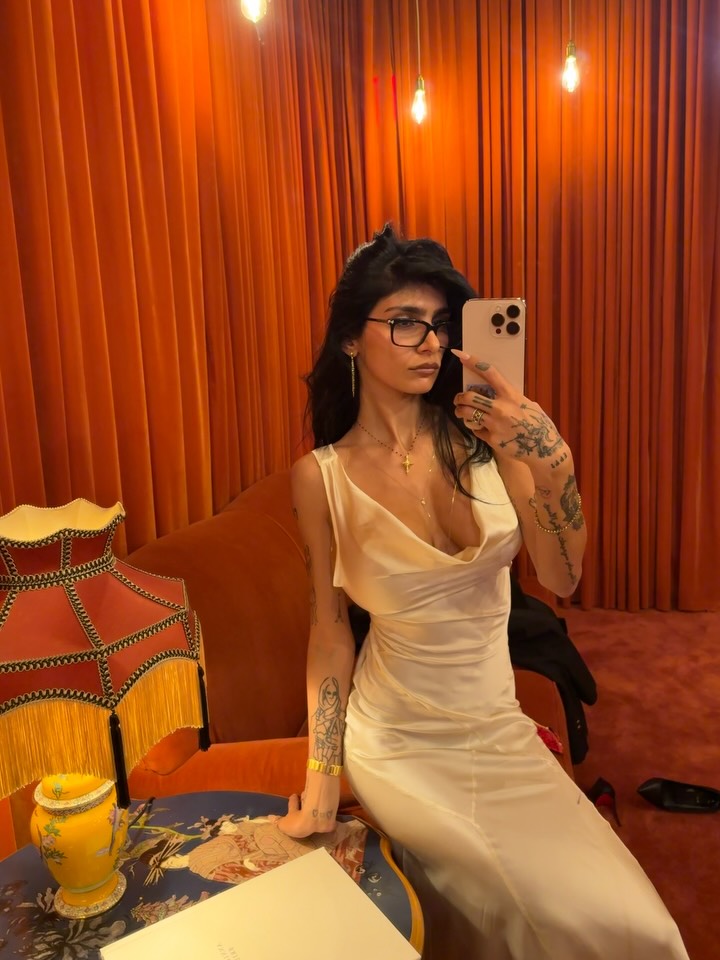Mirror selfie of tattooed OnlyFans creator Mia Khalifa in an elegant white slip dress and glasses inside a room with orange curtains.