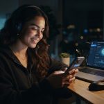 A smiling content creator using a smartphone at night while an open laptop in the background demonstrates how OnlyFans AI bots are used to manage direct messages.