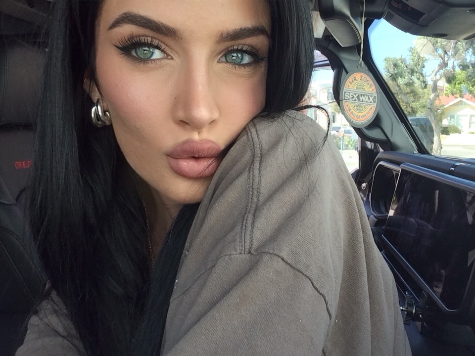 Close-up selfie of California OnlyFans influencer with striking green eyes and full lips, wearing a taupe sweatshirt, inside a vehicle with a "Sex Wax" sticker visible.