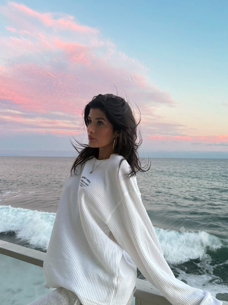 California OnlyFans creator with dark hair, wearing an oversized white sweatshirt, looking out at the Pacific Ocean during a pink and blue sunset.