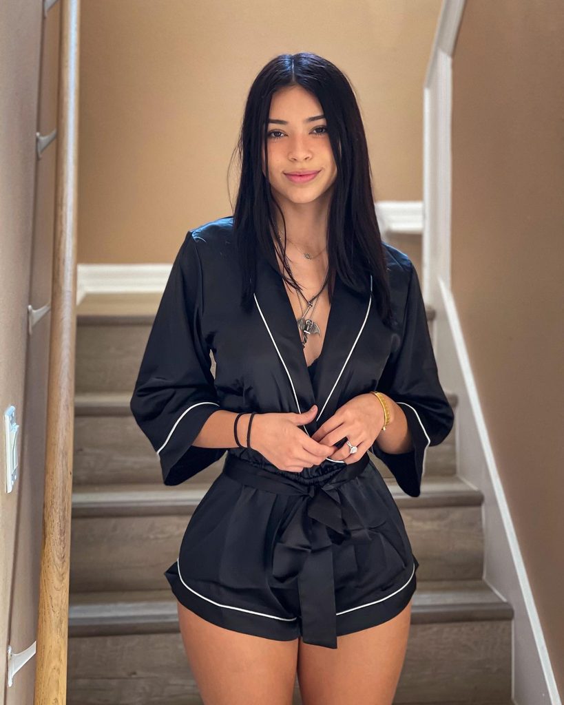 Aishah Sofey standing on stairs in a black satin romper with white piping, smiling at the camera