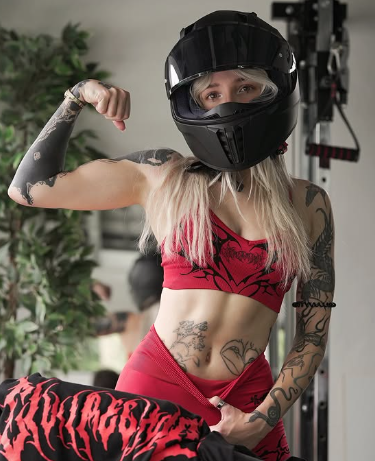 A blonde woman flexing her tattooed arm while wearing a black motorcycle helmet, a red crop top, and red leggings.
