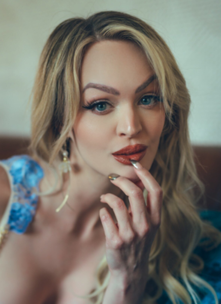 A close-up portrait of a blonde woman with blue eyes and red lipstick, wearing a blue and lace outfit, promoting her Cosplay OnlyFans account.