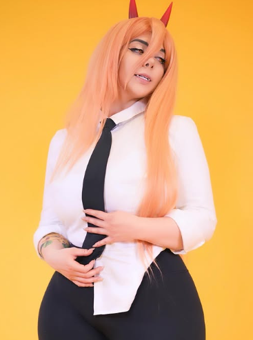 A woman in a Cosplay OnlyFans costume of Power from Chainsaw Man. She wears a white shirt, black tie, black pants, and red horns, standing against a yellow background.