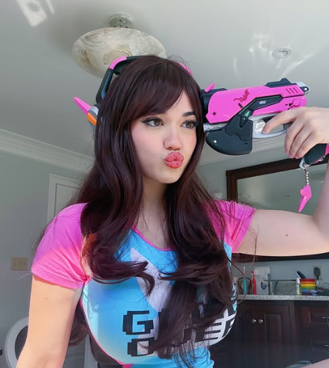 A woman in a D.Va cosplay from Overwatch, wearing a pink and blue shirt and holding a pink prop gun to her head. She has dark hair and is making a kissing face, promoting her Cosplay OnlyFans.