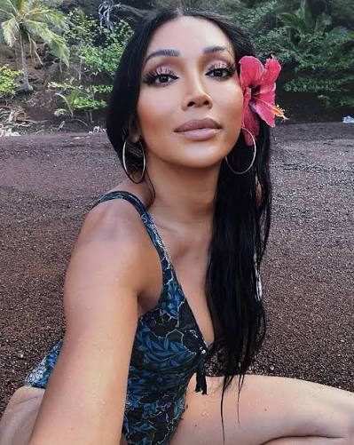 A close-up selfie portrait of popular Asian American OnlyFans creator Yasmin Lee sitting on a beach, wearing a dark patterned swimsuit, gold hoop earrings, and a bright pink hibiscus flower tucked into her long black hair